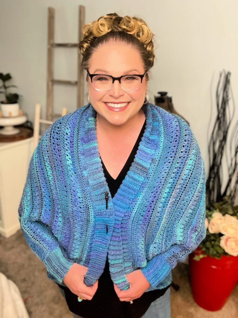 A smiling woman with blonde hair and glasses stands indoors, wearing a blue and purple crochet ruana pattern shawl over a black top. She holds the shawl closed, with flowers and a wooden ladder in the background. -Marly Bird