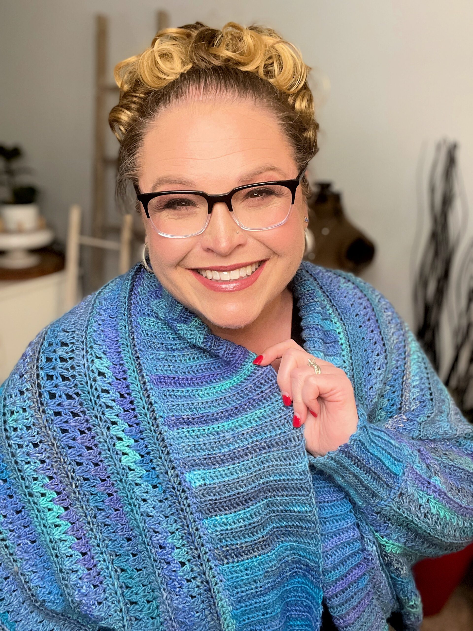 A smiling woman with curly blonde hair, wearing glasses and a colorful oversized crocheted sweater, posing indoors with a soft focus background. You Are Valued Crochet Cocoon Cardigan pattern by Marly Bird