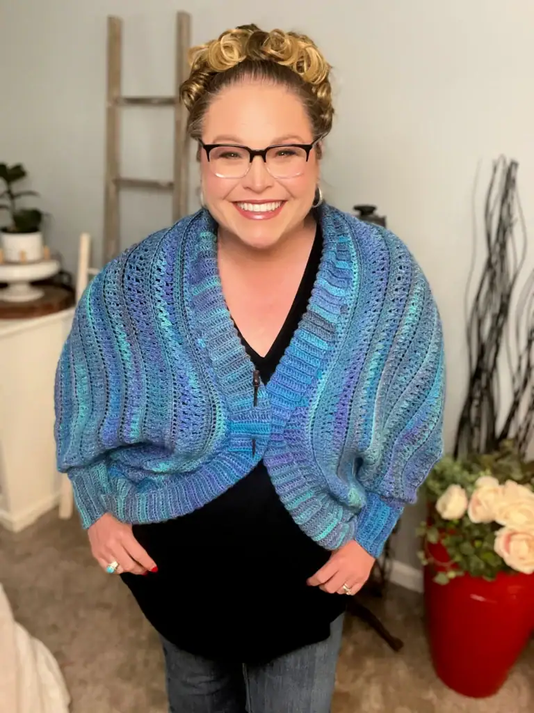 A woman with curly blonde hair and glasses smiles while wearing a blue knitted cardigan over a black top, possibly made from free knitting and crochet patterns. She stands indoors beside a red pot of white flowers and a wooden ladder. -Marly Bird