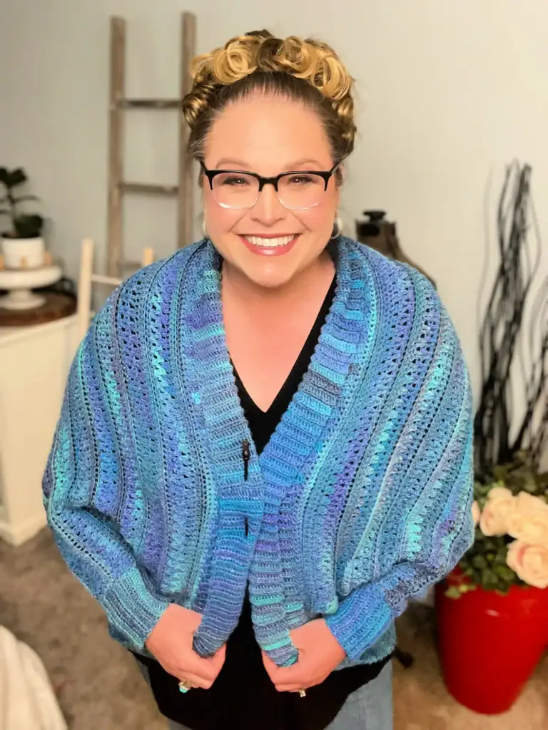 A smiling woman with blond hair and glasses wears a blue and purple crocheted shawl over a black top. She stands indoors in a cozy room with a ladder shelf, a red pot of white flowers, and free knitting and crochet patterns on display. -Marly Bird
