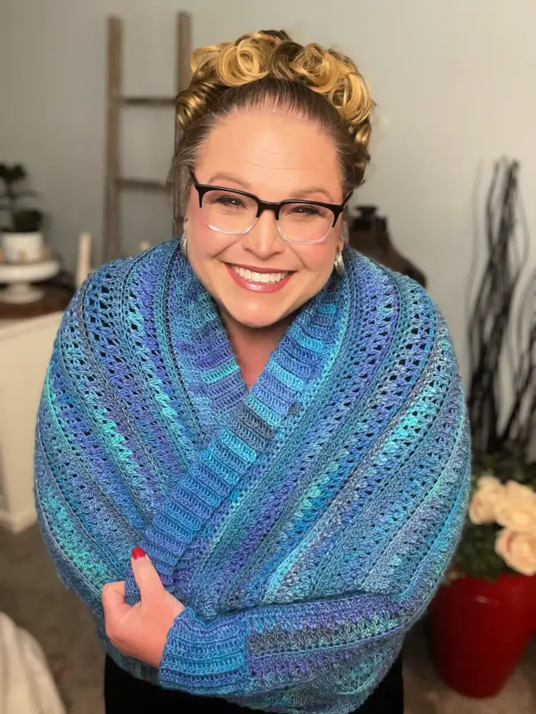 A smiling woman with glasses and curly blonde hair is wrapped in a blue and purple crocheted shawl made from free knitting and crochet patterns. She stands indoors near a small ladder, a vase with twigs, and a red pot of cream-colored flowers. -Marly Bird