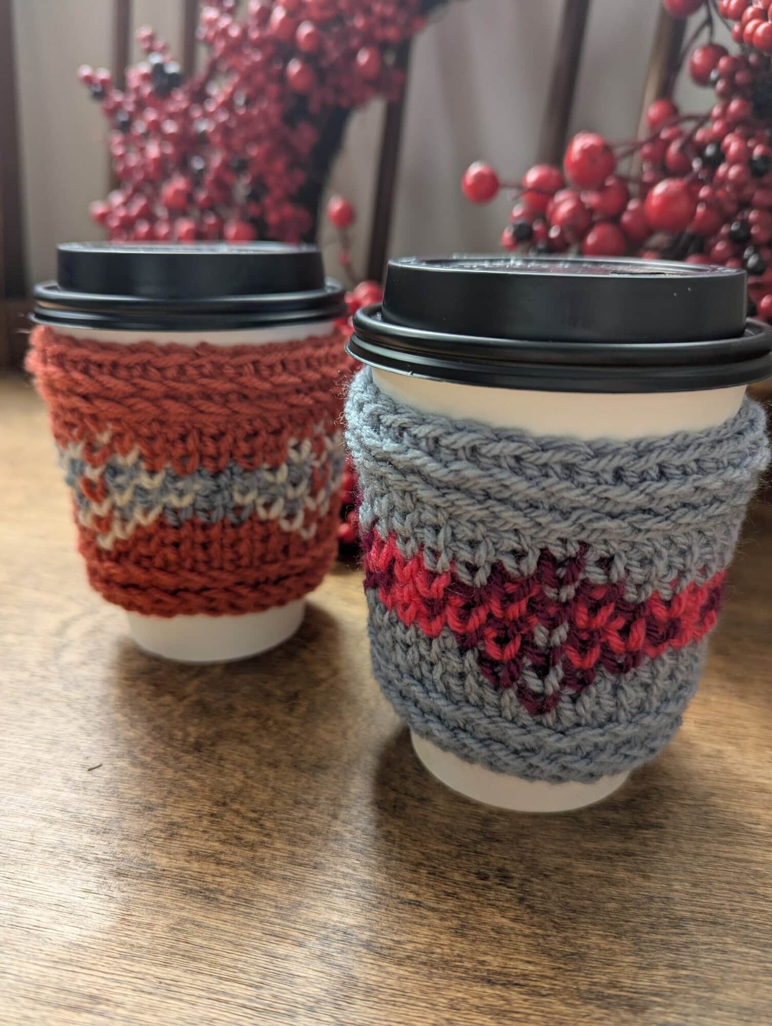 Two coffee cups with black lids feature hand-knitted cup sleeves in blue-pink and red-gray, showcasing textured stitching.