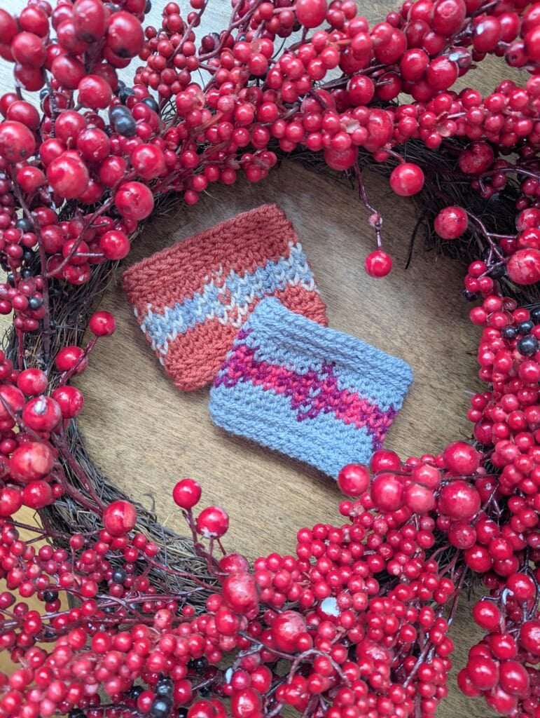 Two small, colorful knit pouches with textured stitches rest on wood, accented by bright red artificial berries and branches.