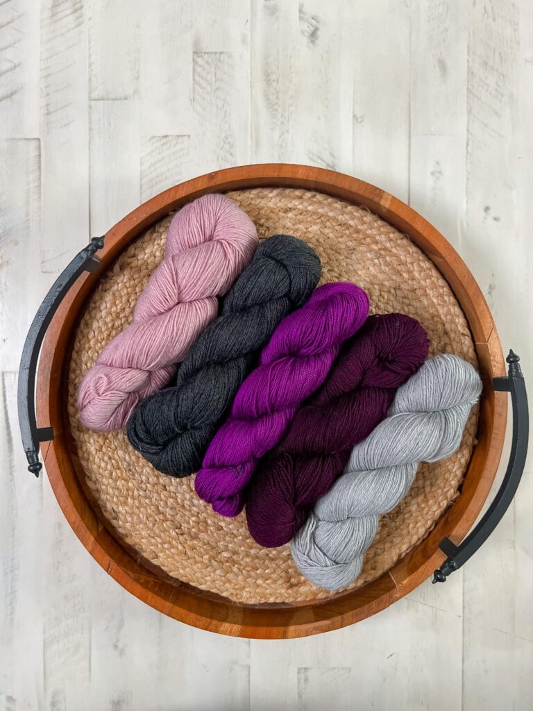 Five skeins of yarn in pink, black, purple, maroon, and gray arranged in a wooden tray, ideal for knit mosaic shawl projects.
