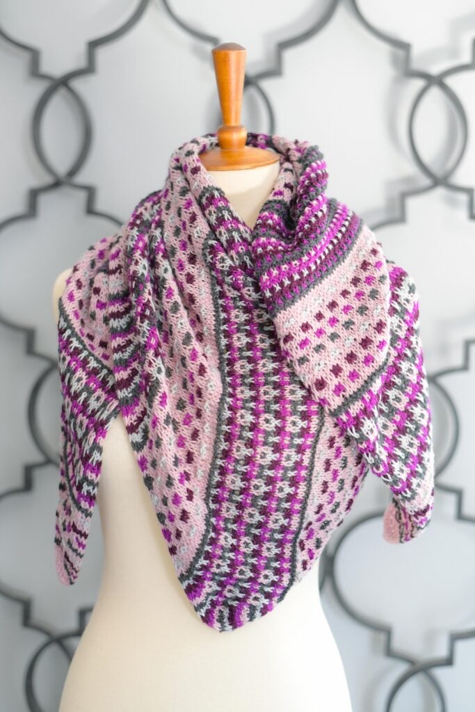 A triangular crochet shawl features mosaic, striped, and geometric stitches in pinks, purples, white, and gray on a mannequin.