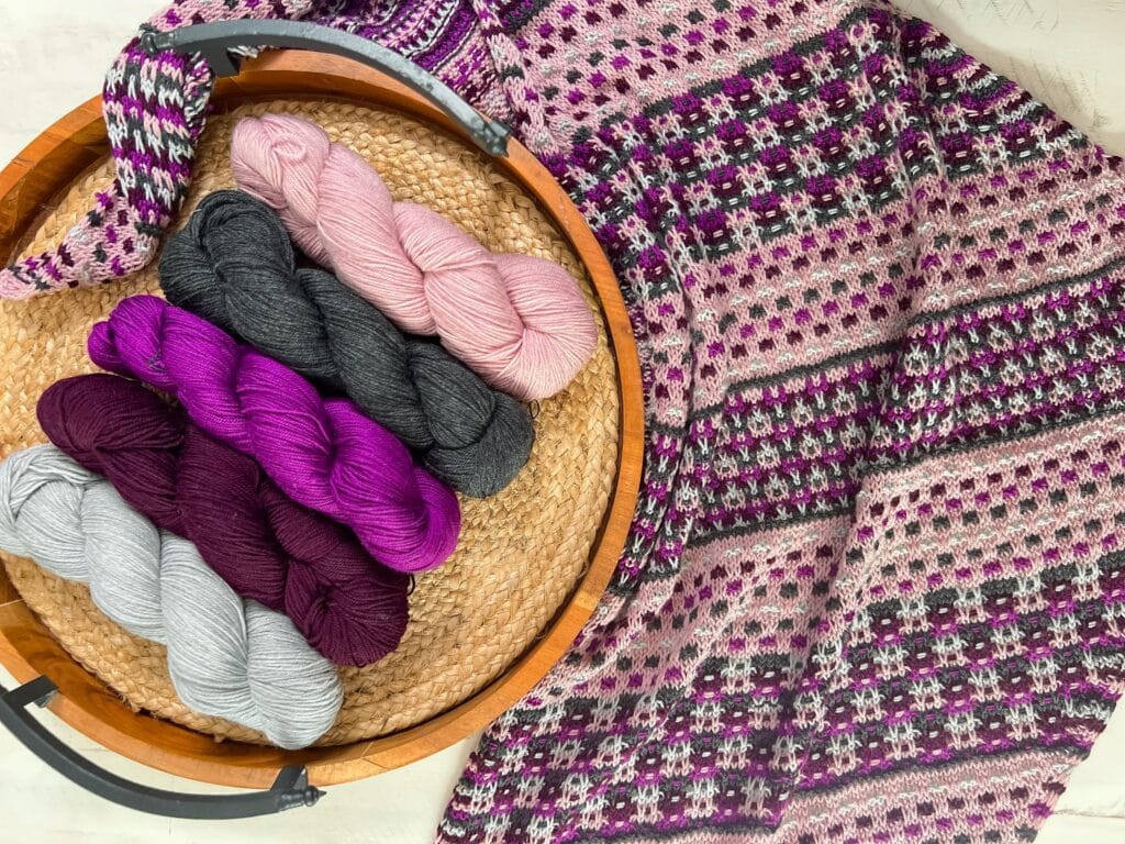 Slip stitch mosaic knitting detail in five contrasting colors. A knit mosaic shawl with geometric patterns and rich pink, purple, gray, and burgundy tones, displayed beside matching yarn skeins.