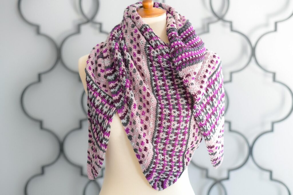 Large knit mosaic shawl in pink, purple, white, and gray with geometric slip stitch pattern, draped on a mannequin’s shoulders.