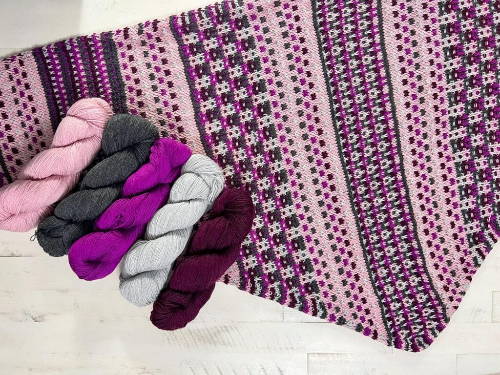 Fingering weight knit mosaic triangle shawl with slip stitch pattern laying flat with Pink, purple, gray, and white yarn laying on top of the shawl and white wood background.