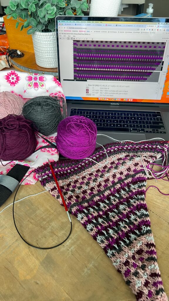 working view of a corner to corner knit shawl construction at the start of a project. A textured crochet triangle shawl lies on a table with colorful yarn, a floral project bag, and a laptop showing a stitch chart. 