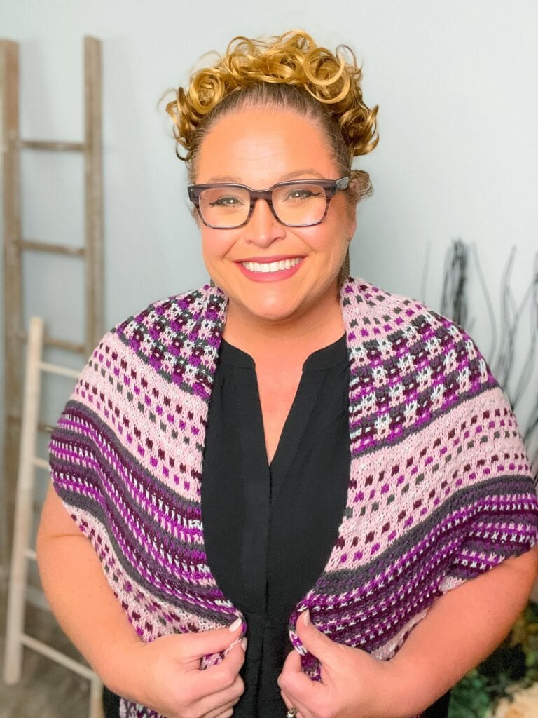 The Slip Stitch shawl in purple, pink, and white shows intricate stitch detail; modeled over a black top indoors.