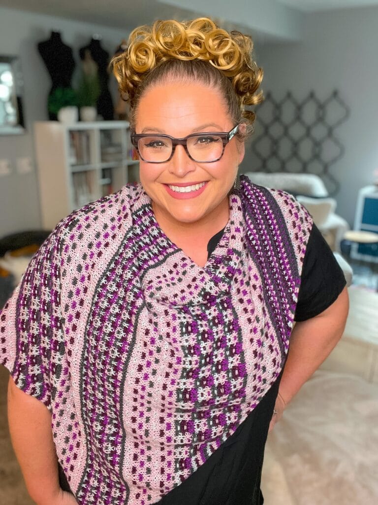 Woman models a purple patterned mosaic knit shawl, showing stitch texture. Cozy indoor setting with shelves behind her.