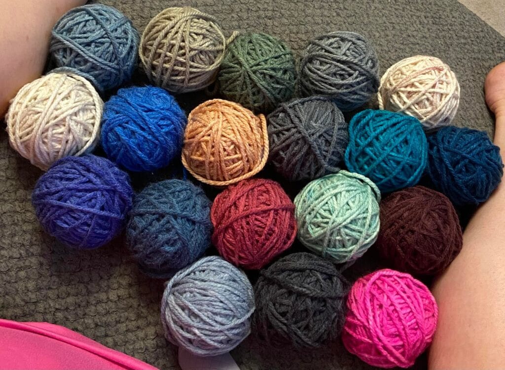 Balls of yarn in blue, gray, green, red, brown, pink, and cream are laid on a textured gray carpet beside two bare legs.