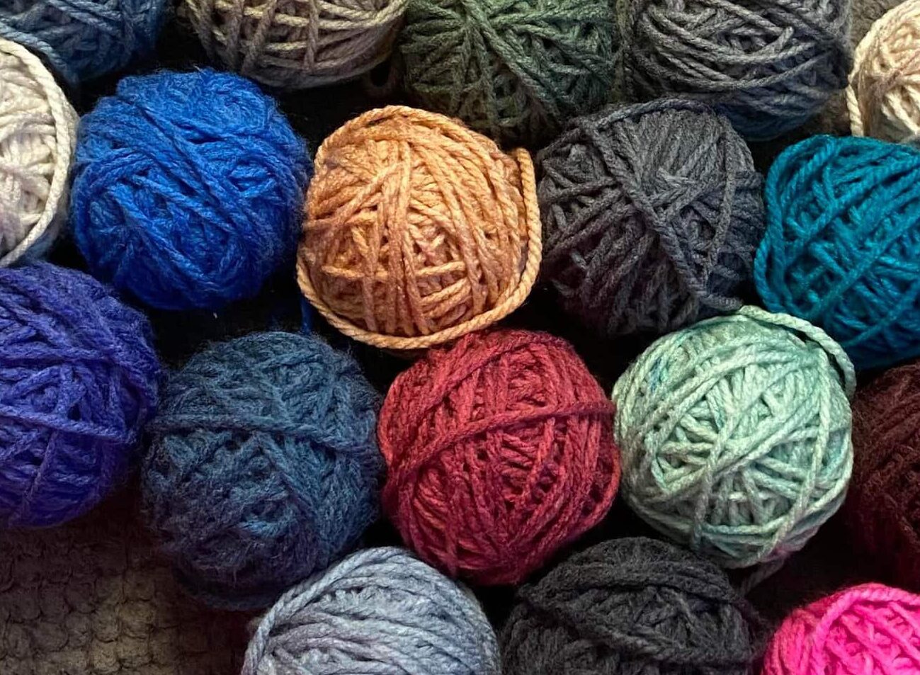 Balls of yarn in blue, gray, green, red, brown, pink, and cream are laid on a textured gray carpet beside two bare legs.