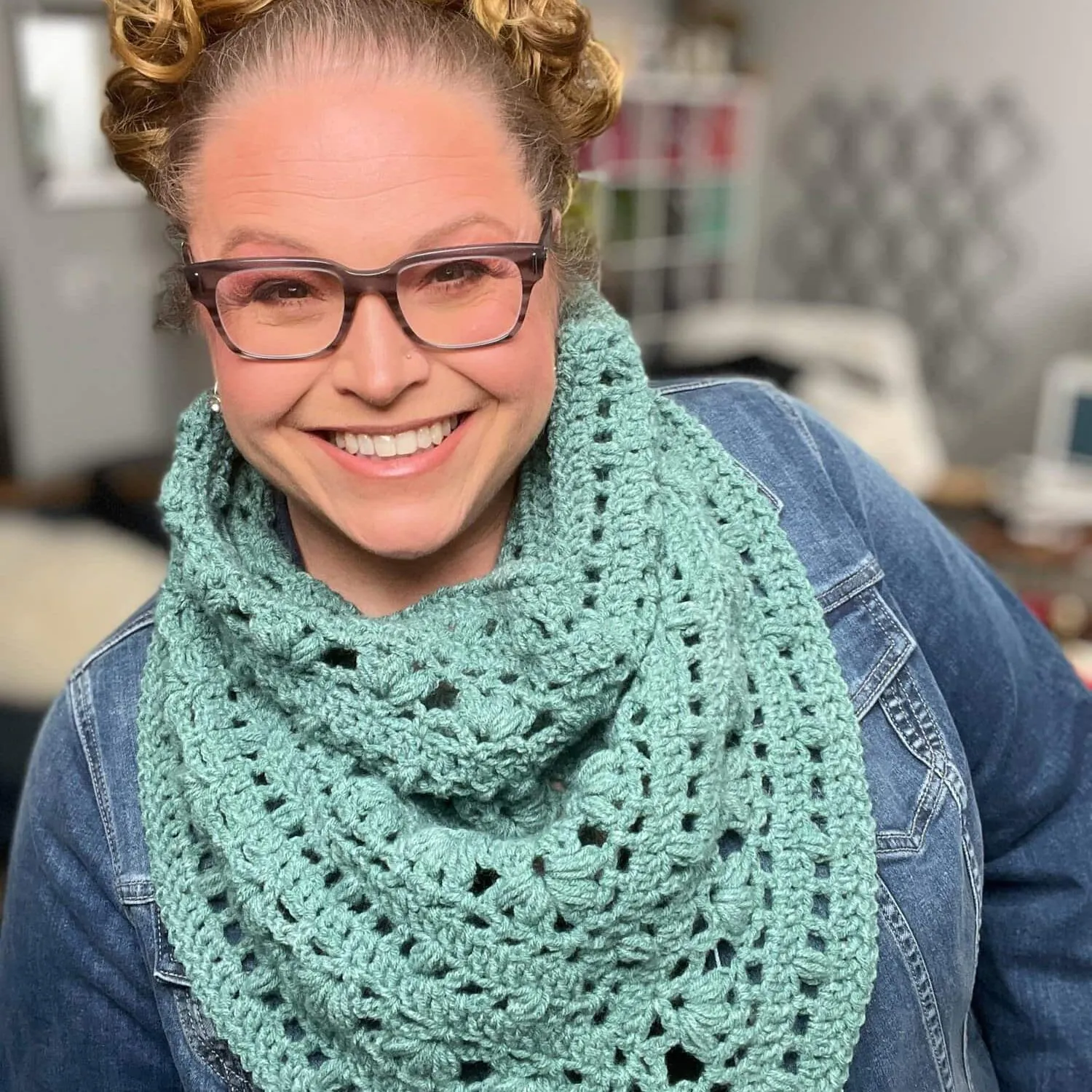 Woman models a light teal crocheted shawl with visible textured stitches, layered over a denim jacket in a well-lit room.