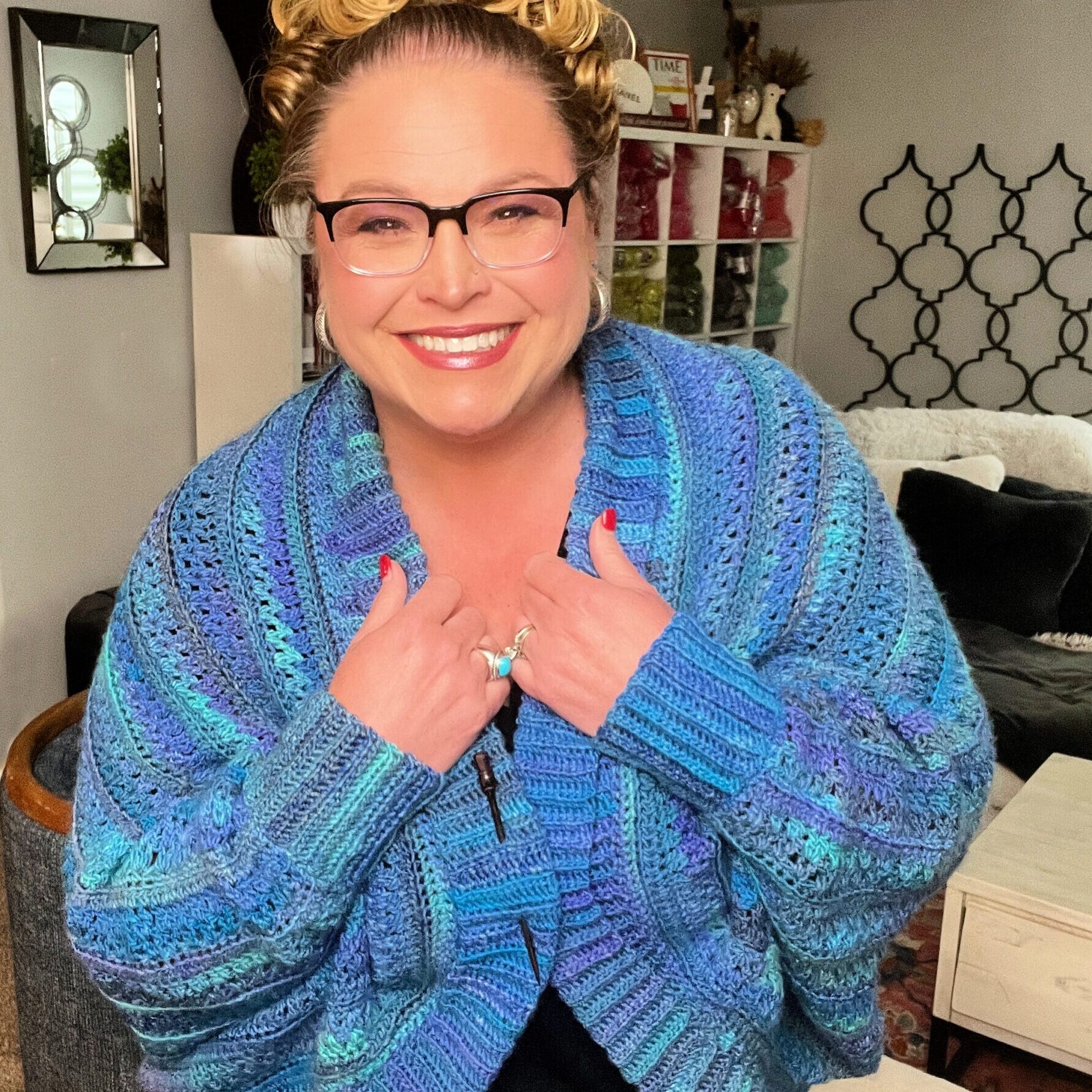 A smiling woman with curly updo hair, wearing glasses, beams as she models the You Are Valued Crochet Cardigan in a vivid blue, showcasing the crochet texture against a cozy home backdrop - You Are Valued Crochet Cocoon Cardigan Marly Bird.