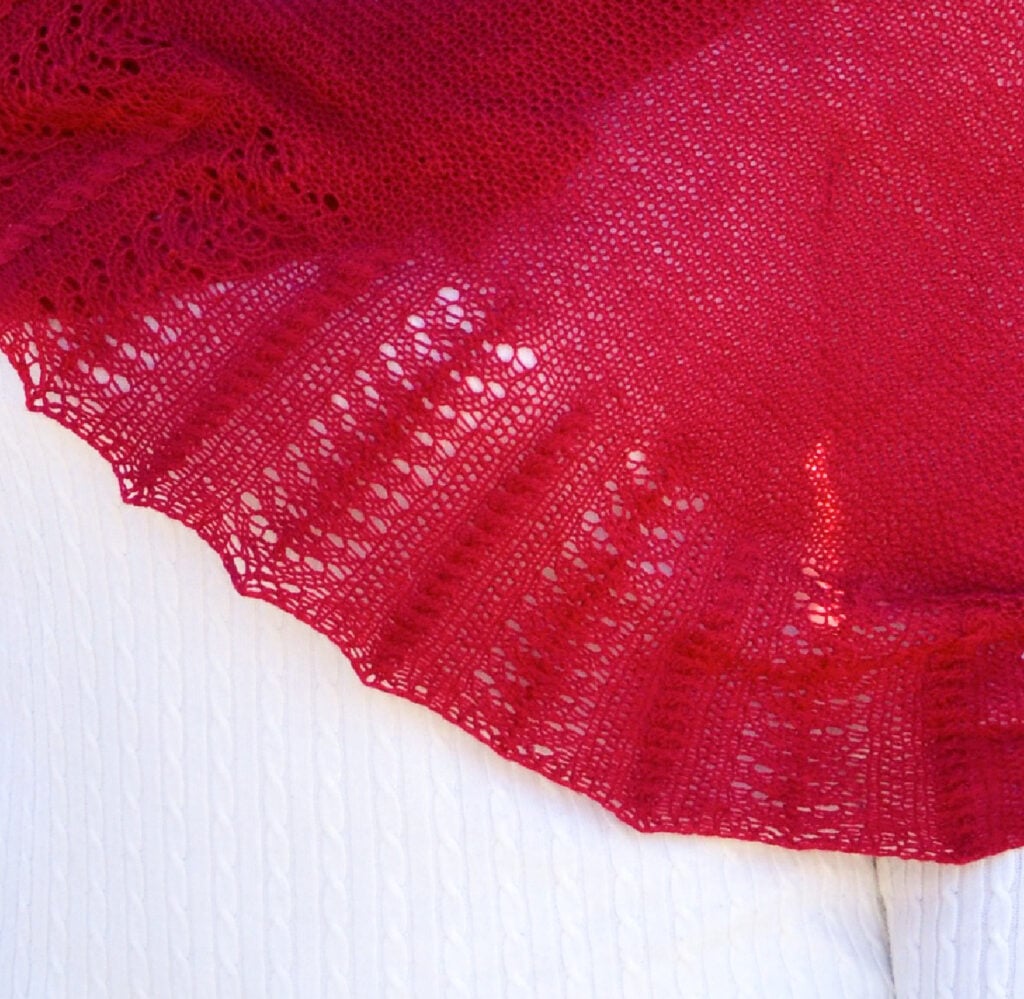 Red knit shawl with lacy scalloped edges, draped over white textured fabric, showing stitch detail and texture.