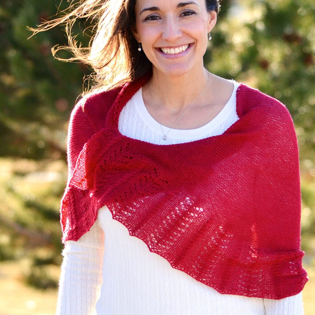 A woman models a red knit Outlander shawl with textured stitches, draped over her white sweater, outdoors among green trees.