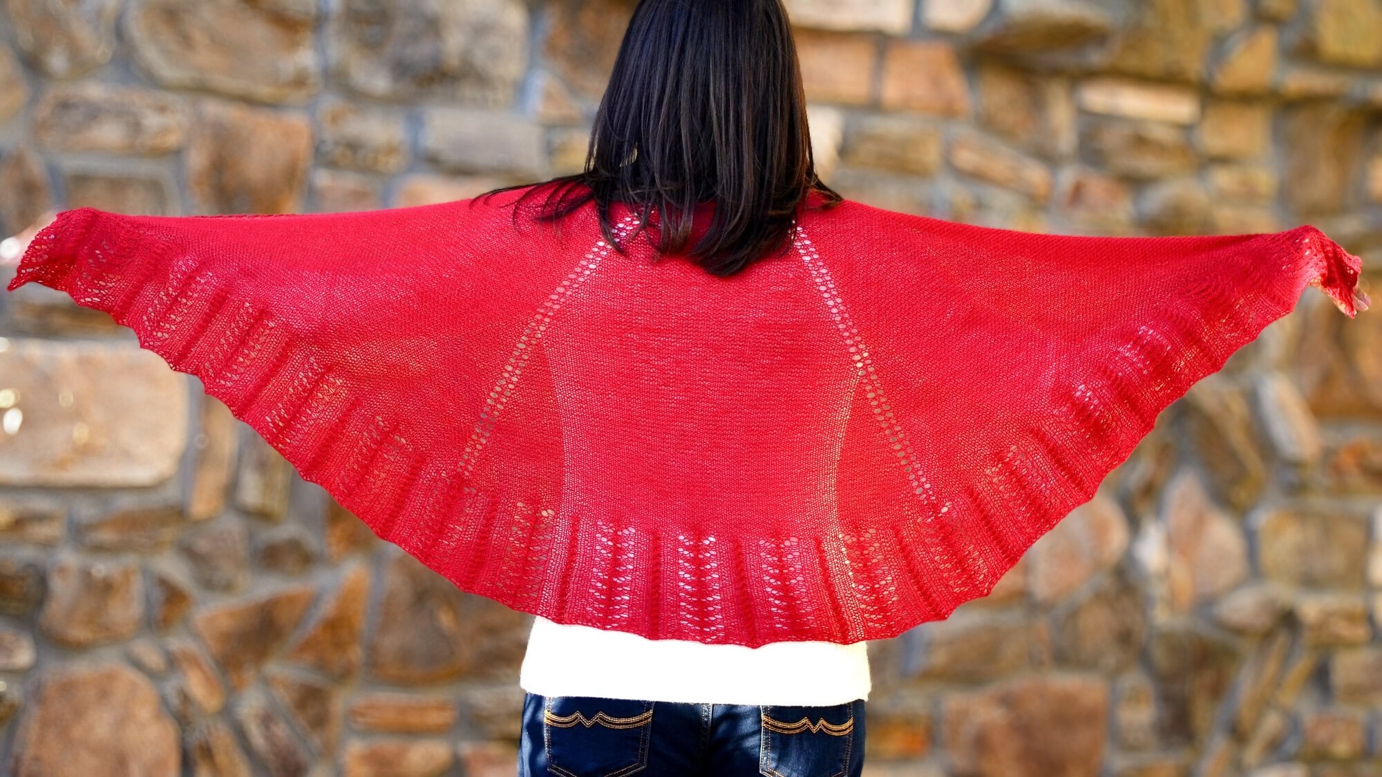 A bright red knit Outlander shawl with visible stitch detail is spread wide across the back of a person facing a stone wall.