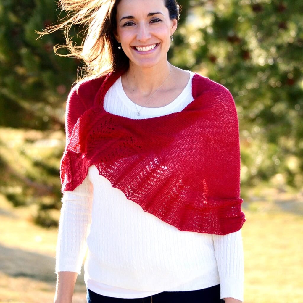 A woman models a red Outlander knit shawl with textured stitches over a white sweater, outdoors in sunlight.