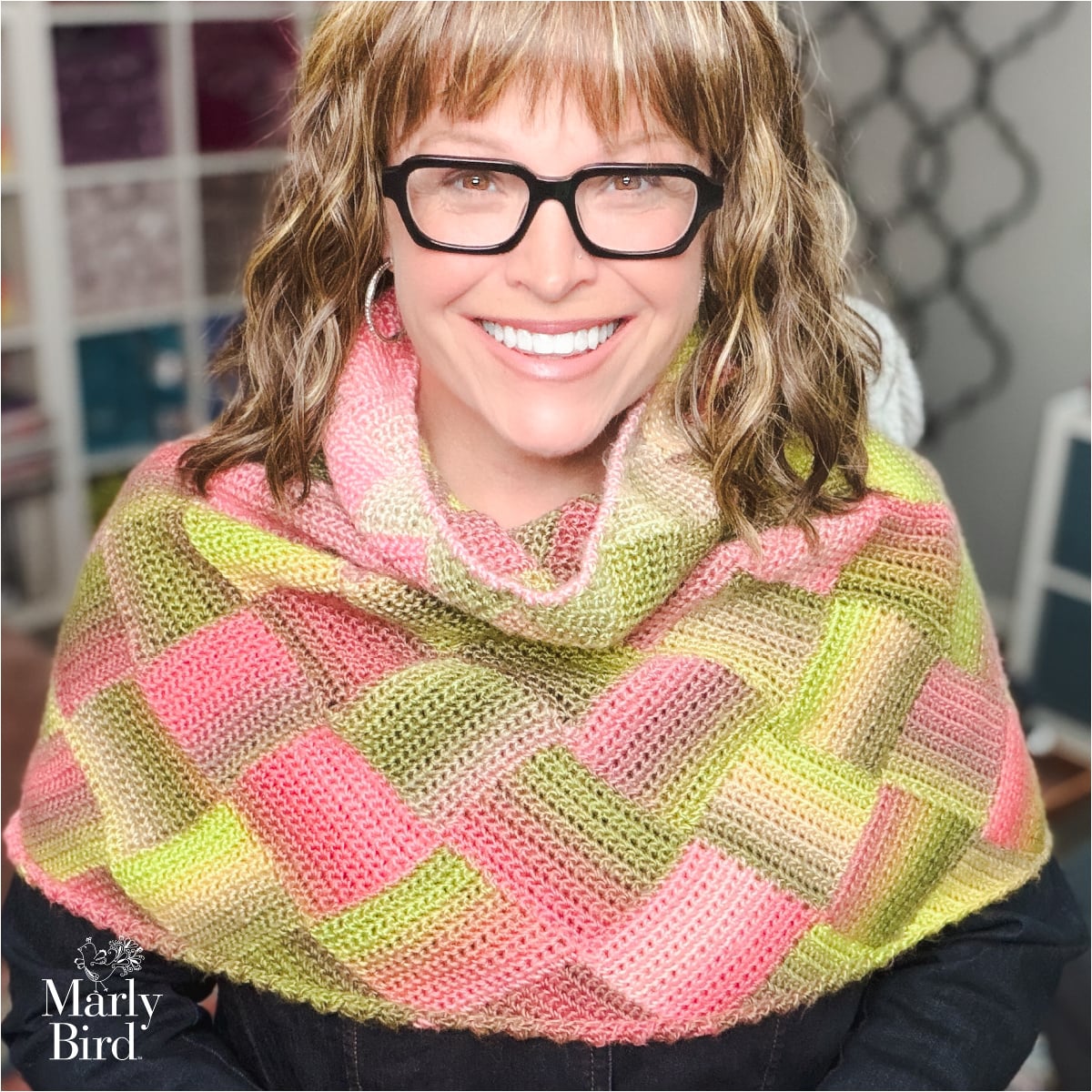 A smiling woman models a pink, green, and beige crochet entrelac shawl; shelves of yarn sit against a gray patterned wall behind her.