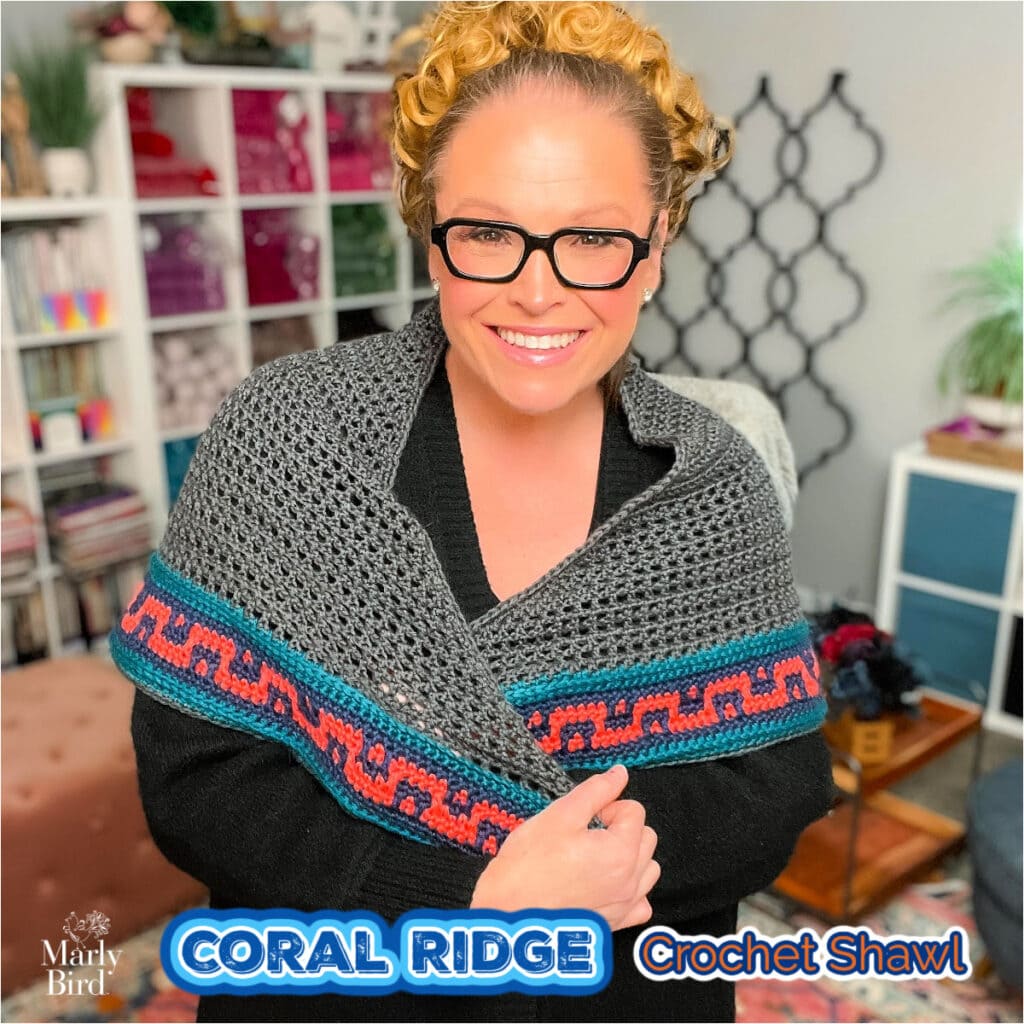 Coral Ridge free crochet triangle shawl pattern — handmade crochet shawl in gray with blue and coral colorwork border, styled in a cozy craft room