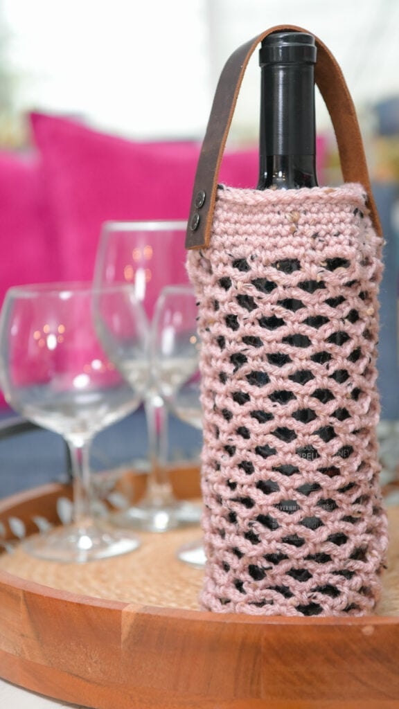 crochet wine cozy handmade hostess gift idea. Pink crocheted wine holder with leather handle on tray, showing textured stitches; three glasses and bright pink cushions nearby.