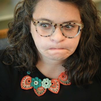 A woman with curly hair and glasses, wearing a black top with a colorful crochet necklace, looks directly at the camera with a slight frown. -Marly Bird