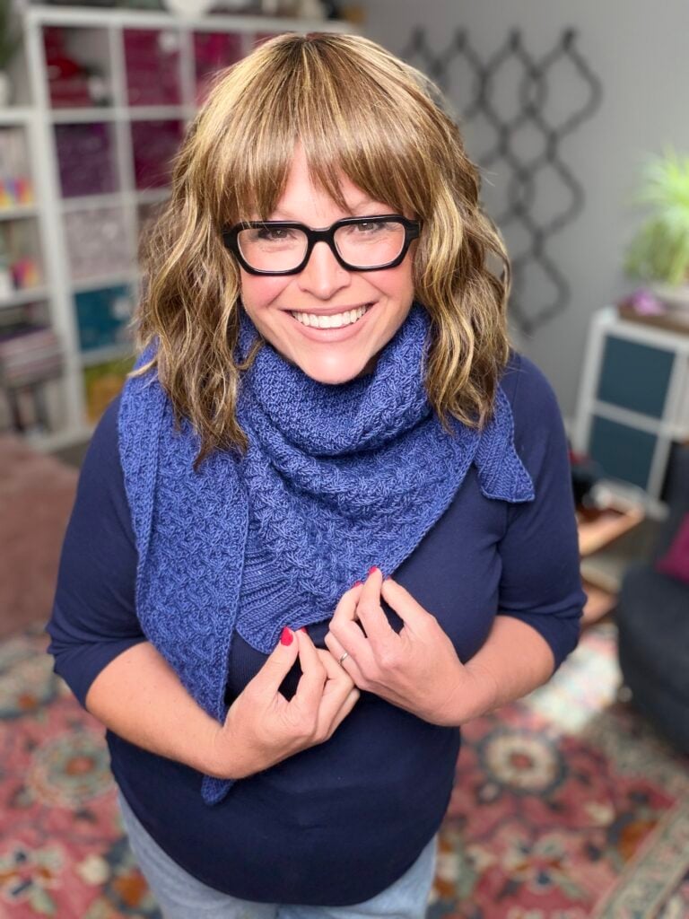 Woman modeling a blue textured triangle shawl over a navy shirt, showing stitch detail in a bright, cozy room.
