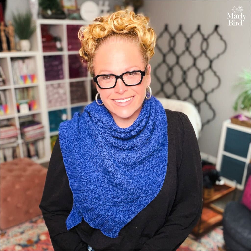 A woman models a textured triangle knit shawl over black, in a cozy room with bookshelves and yarn in the background.