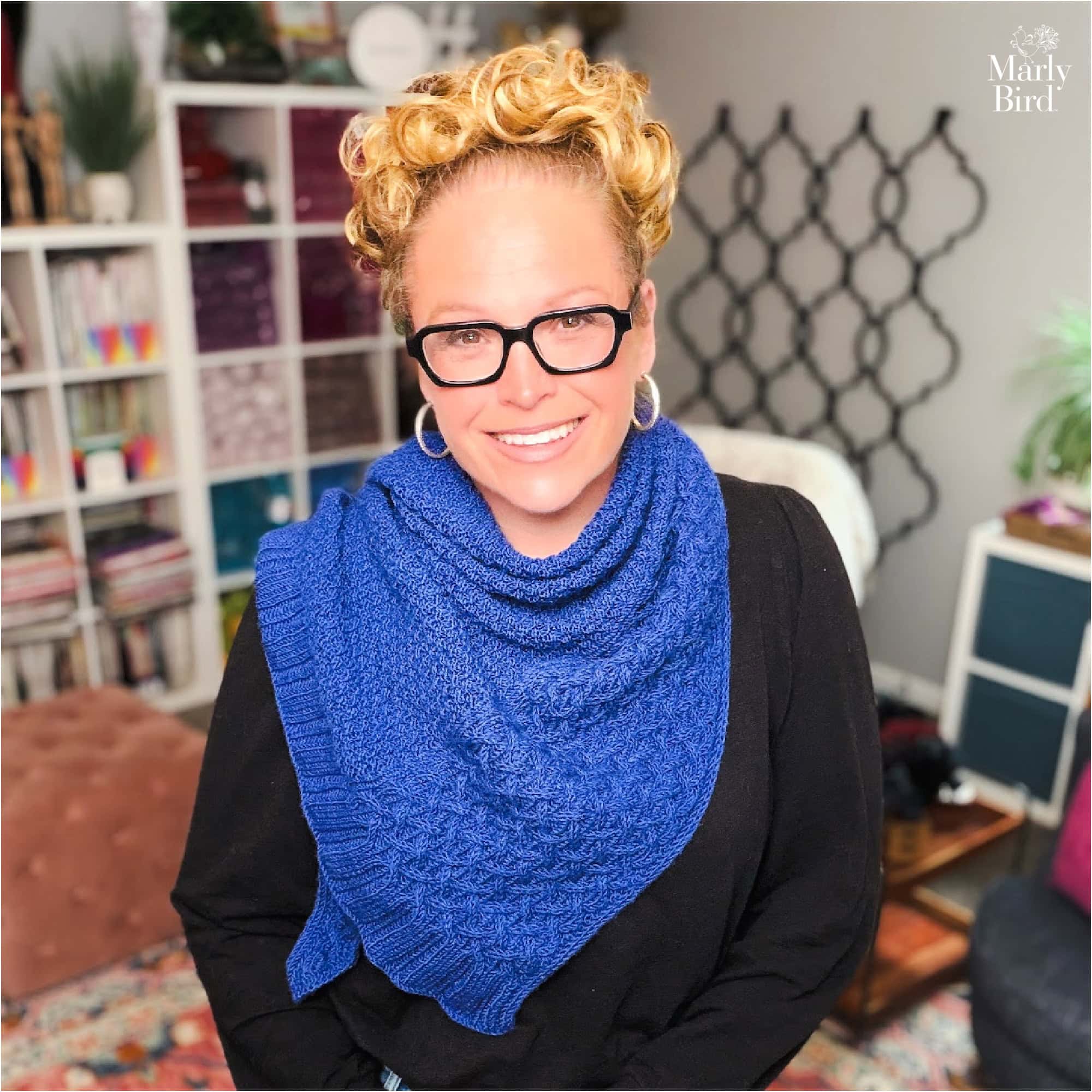 A woman models a textured triangle knit shawl over black, in a cozy room with bookshelves and yarn in the background.