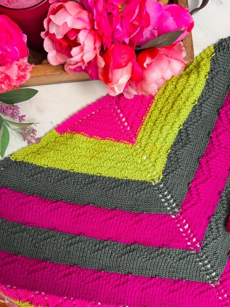 Striped shawl in pink, lime green, and gray yarn shows intricate stitch detail. Shawl is laid beside a tray of pink flowers.