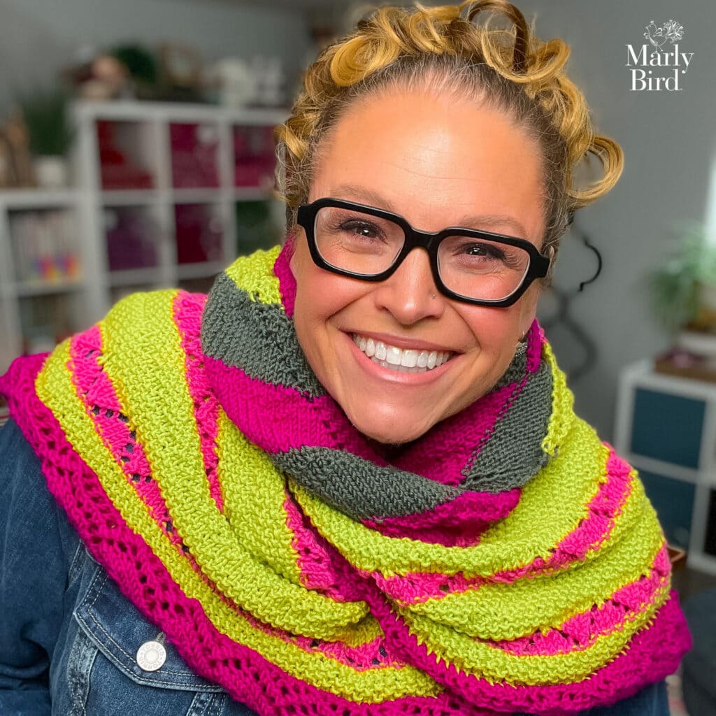 Person models a vibrant pink, green, and gray knitted shawl with textured stitch detail; yarn shelves and Marly Bird logo behind.
