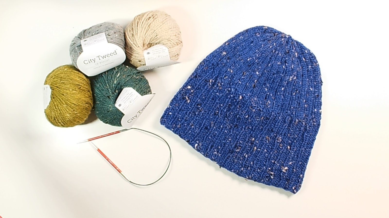 On a table are four balls of the City Tweed DK Yarn in different colors, a pair of 16 inch size 7/4.5mm knitting needles, and the Every Day Knit Hat laying flat with the brim folded down - Marly Bird