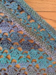 Lacy crochet fabric in blue, green, and purple with a floral pattern and soft texture, displayed on a wooden surface.