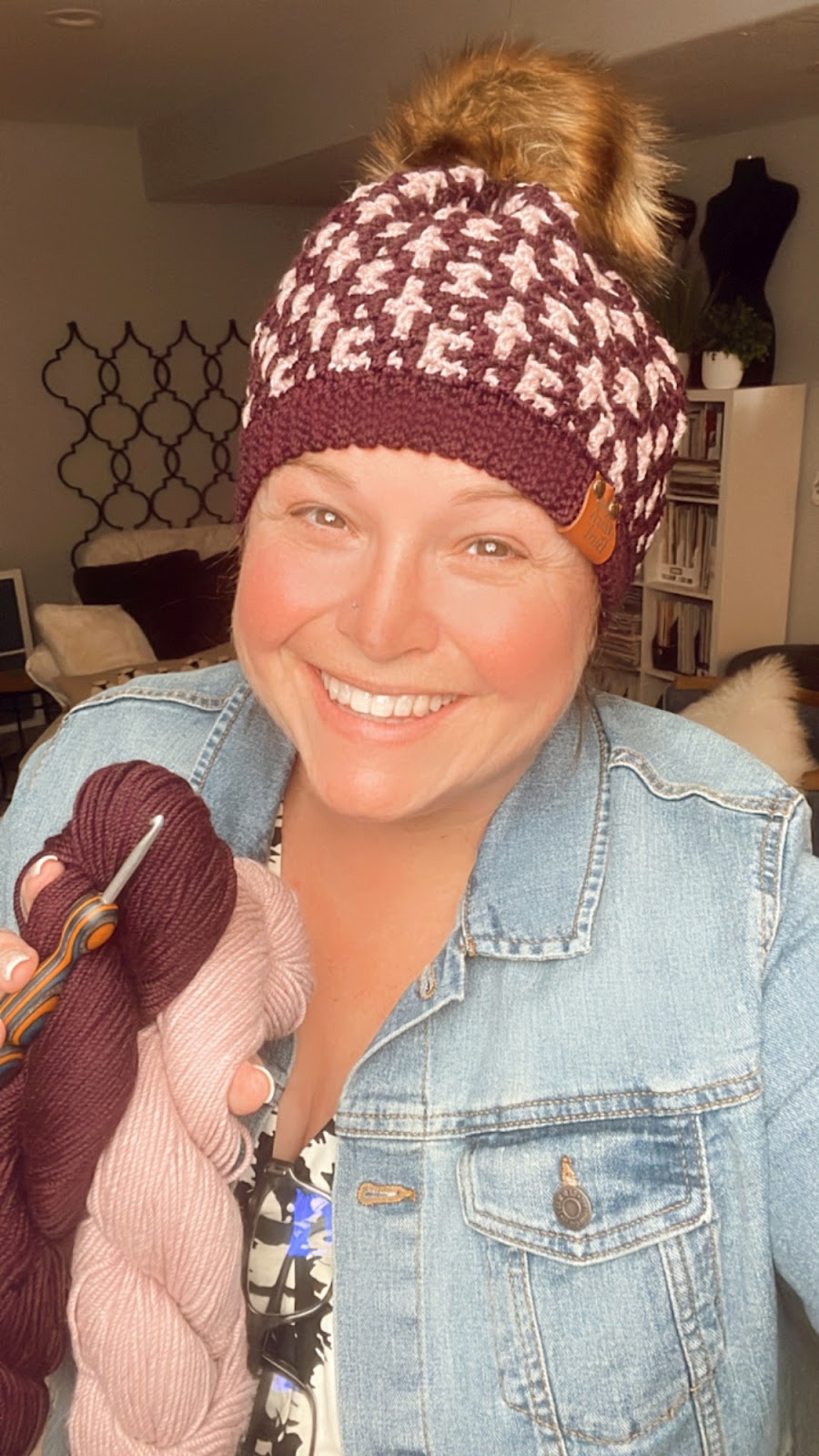 A person smiles warmly while holding two skeins of yarn and a crochet hook. They are wearing a knitted hat with a pom-pom and a denim jacket. The background appears to be a cozy living room with a patterned accent wall and various decorations.