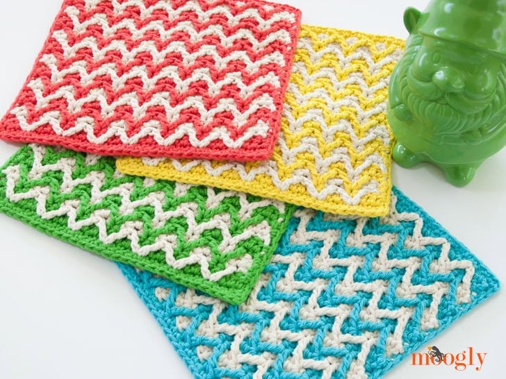 Stacked crochet dishcloths in red, yellow, green, and blue with white zigzag stitch detail, beside a green gnome figurine.
