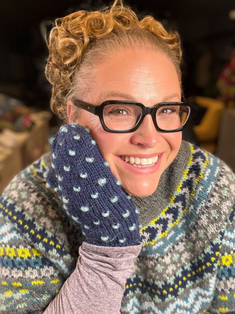 A smiling person with curly hair and glasses wears a colorful patterned sweater and holds their cheek with a navy blue mitten featuring white polka dots—perfect inspiration for your next Andes knit mittens or free knitting pattern project. Bright light highlights their cheerful expression. -Marly Bird