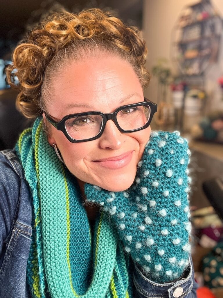 A person with curly hair and glasses smiles at the camera, wearing a blue and green knit scarf and a fleece-lined crochet mitten. The background is softly blurred, showing various household items—perfect inspiration for a Marly Bird fan. -Marly Bird