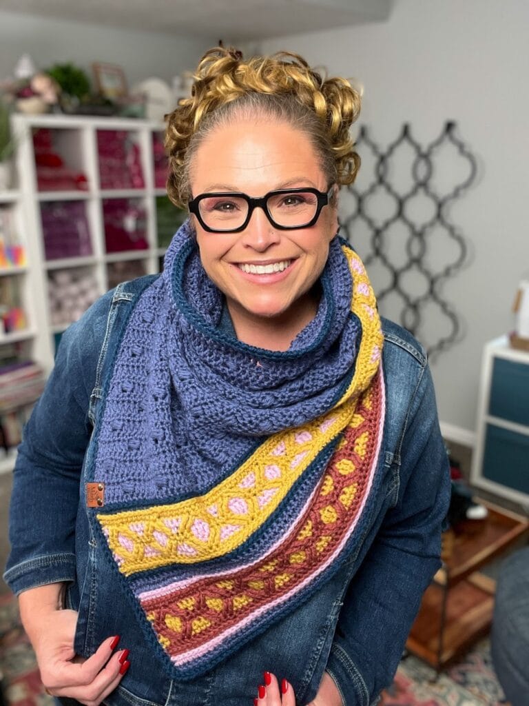 A smiling woman with curly blonde hair and glasses wears a denim jacket and a colorful, crocheted Ridgeway Crochet Cowl in blue, yellow, pink, and maroon patterns, standing in a cozy, yarn-filled room. -Marly Bird