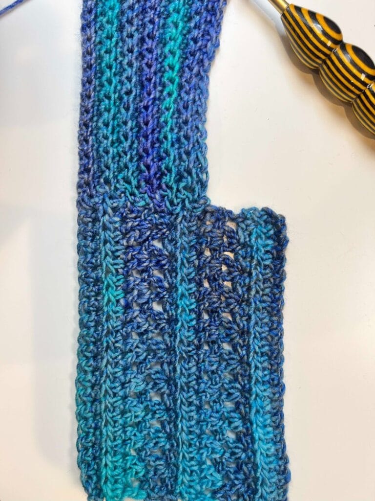 A crocheted piece in shades of blue and teal with a ribbed texture and a rectangular section branching out to one side, ideal as part of a Crochet Cocoon Cardigan, sits next to a crochet hook with a yellow and black striped handle. -Marly Bird