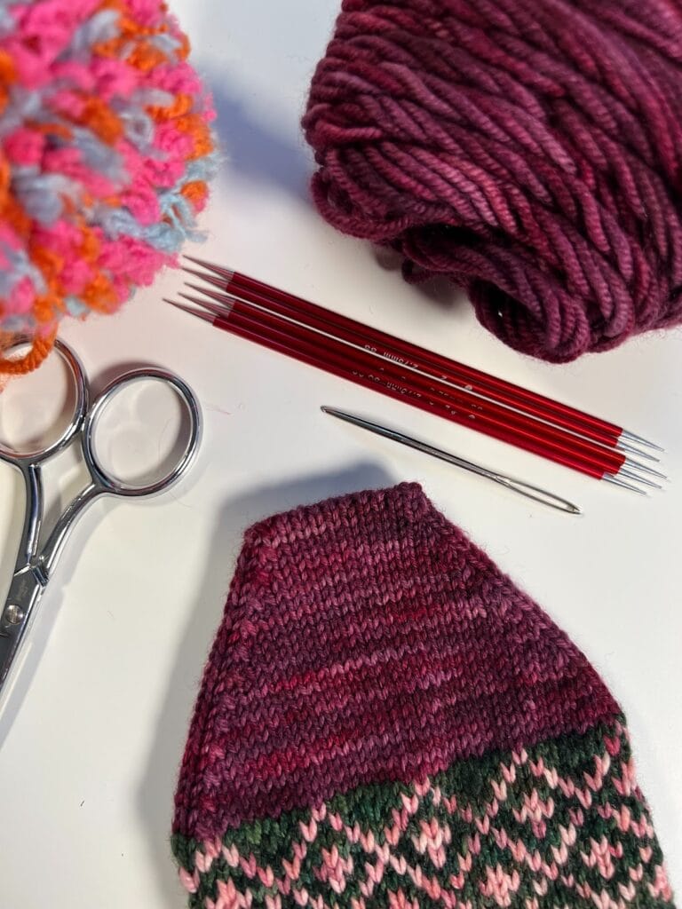A close-up of knitting supplies: a pair of scissors, a large ball of pink yarn, red double-pointed knitting needles, the Heartstrings knit sock pattern, a metal darning needle, and a partially finished knit piece with pink and green patterns. -Marly Bird