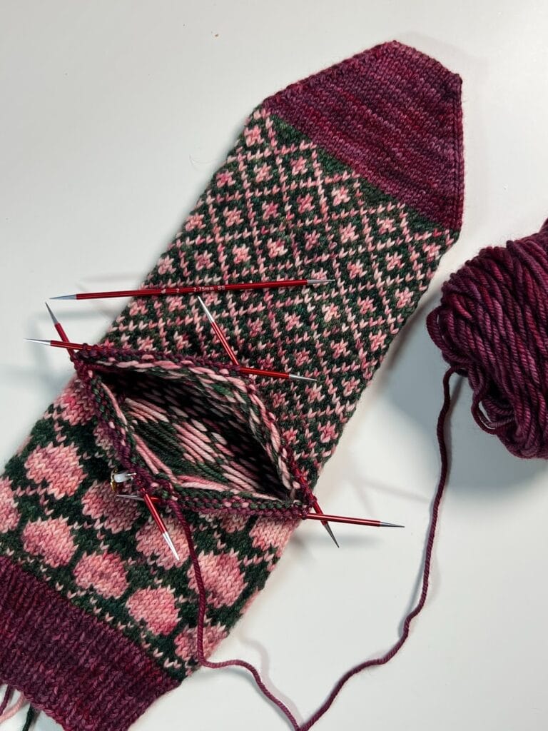 A partially knitted mitten with intricate pink and green patterns, knitting needles inserted, and a ball of maroon yarn attached—evoking the cozy charm of the Heartstrings knit sock pattern—rests on a white surface. -Marly Bird