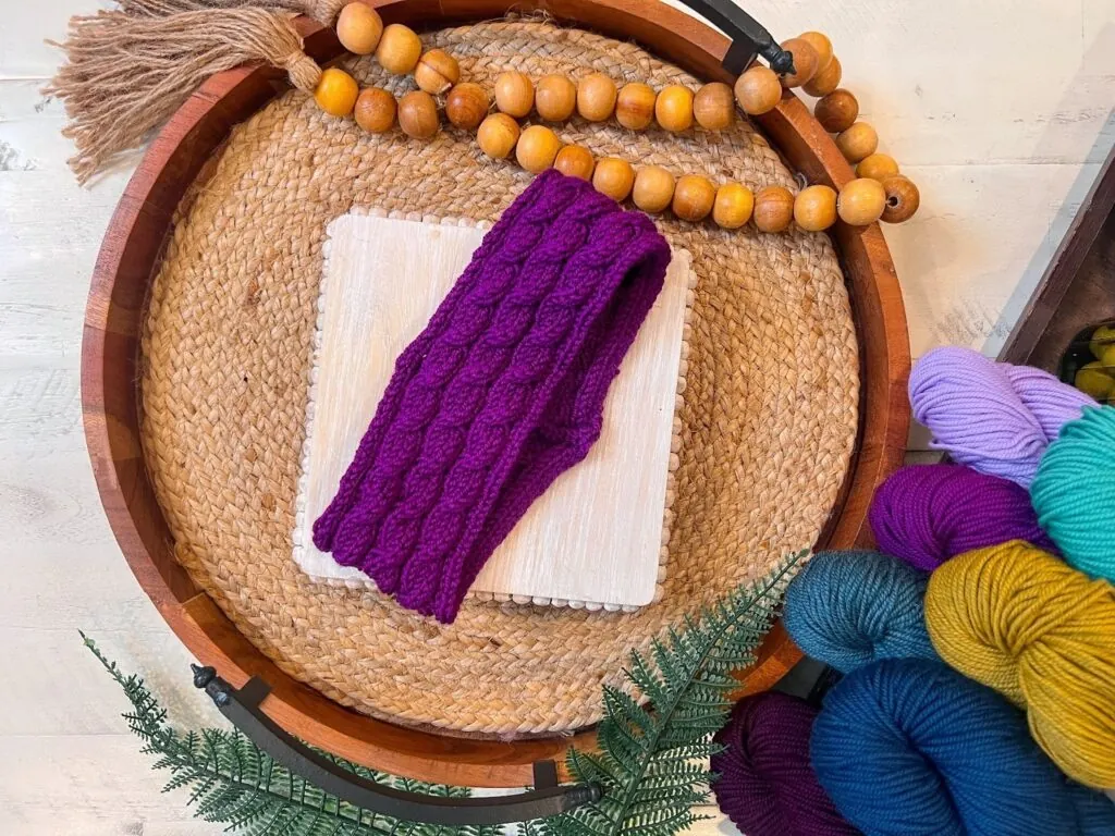 A purple Triple Twist knit ear warmer rests on a square white coaster atop a round woven tray, decorated with wooden beads. Colorful yarn skeins and green fern leaves are visible nearby, perfect for your next free knit pattern project. -Marly Bird