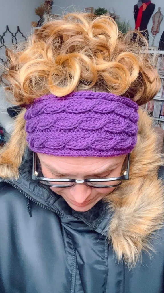 A person with curly, light brown hair wears a purple Knit Ear Warmer and glasses, looking down. They are dressed in a black coat with a fur-trimmed hood. The background features indoor furnishings and shelves. -Marly Bird