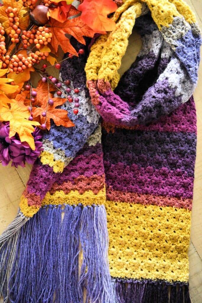 A colorful crocheted scarf in shades of yellow, purple, burgundy, and gray with fringe, lies next to autumn leaves and berries on a light wood surface. -Marly Bird