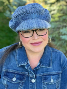 A woman wearing glasses, a blue knit beret made from free knitting and crochet patterns, hoop earrings, and a denim jacket smiles outdoors with greenery in the background. -Marly Bird