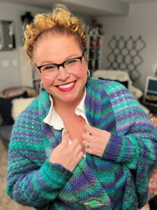 A smiling person with curly blonde hair and glasses wears a colorful blue and green sweater—made using free knitting and crochet patterns—over a white shirt, standing indoors in a cozy, well-lit room. -Marly Bird