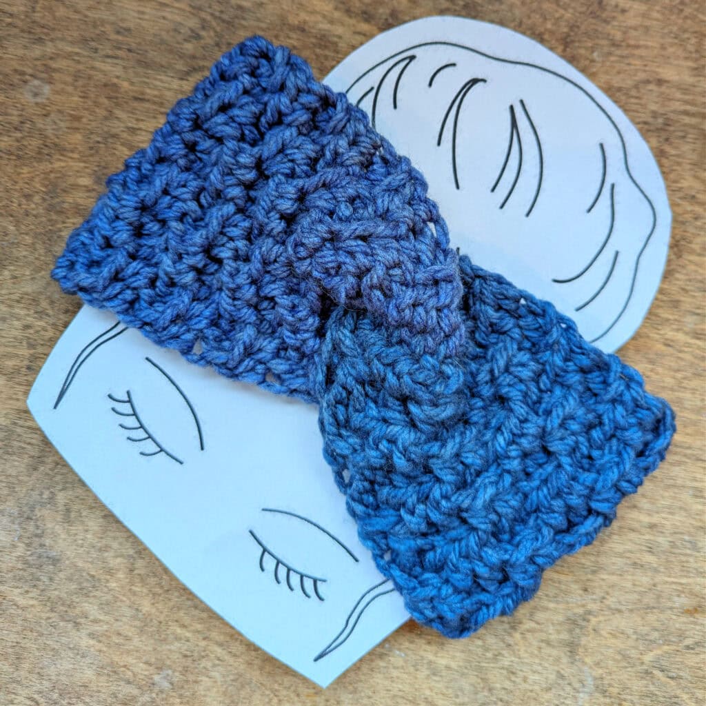 A blue crocheted bow, crafted from a free crochet headband pattern, rests gently atop two illustrated paper faces with closed eyes, drawn on a wooden surface. -Marly Bird