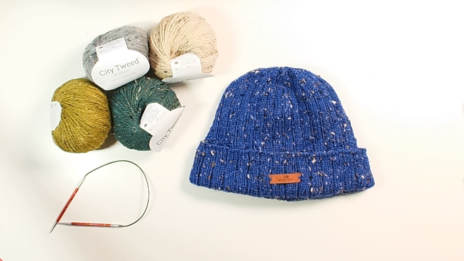 On a table are four balls of the City Tweed DK Yarn in different colors, a pair of 16 inch size 7/4.5mm knitting needles, and the Every Day Knit Hat laying flat with the brim folded up - Marly Bird