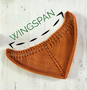 Orange knitted triangle shawl on a white surface; top edge marked with “WINGSPAN” in green and black dashed lines.